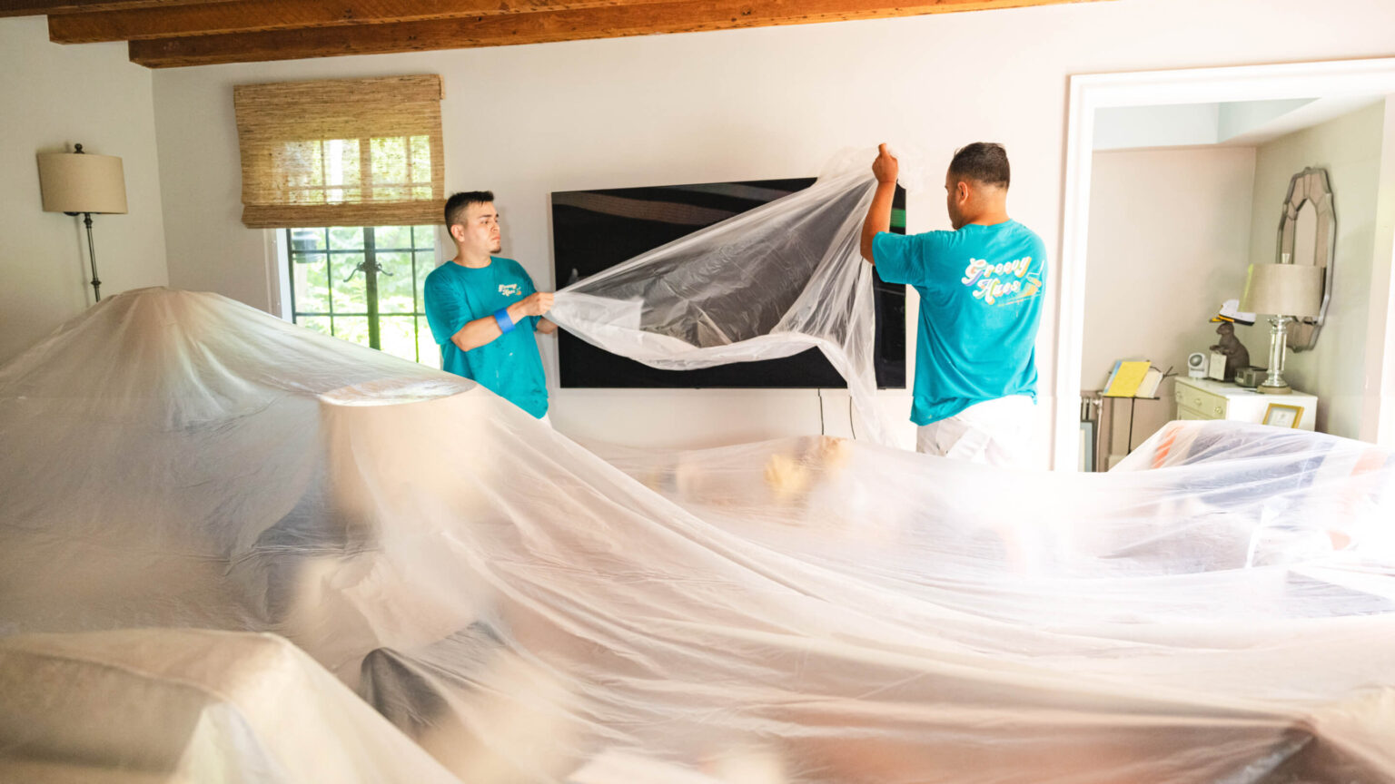 Groovy Hues Painting of Philadelphia crew members in branded teal shirts draping protective plastic sheeting over a client's living room furniture before painting